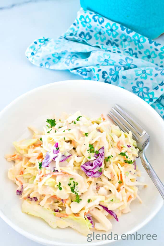 Best Easy Coleslaw Recipe served in a white bowl on a marble countertop. A blue and white print fabric napkin is in the background.