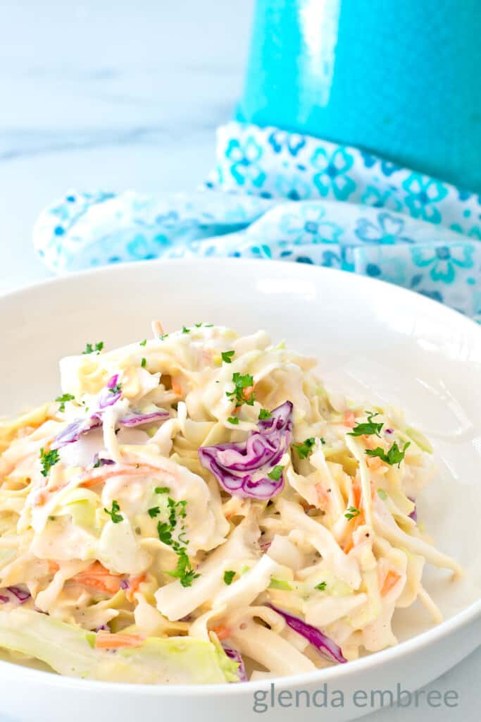 Best Easy Coleslaw Recipe served in a white bowl on a marble countertop. A blue and white print fabric napkin is in the background.