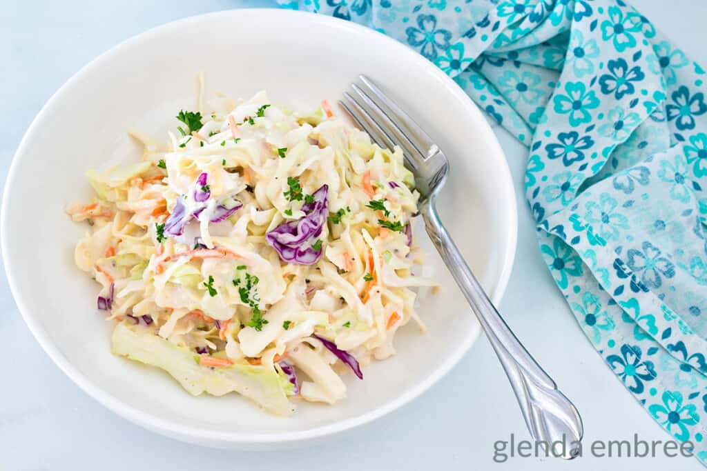 Best Easy Coleslaw Recipe served in a white bowl on a marble countertop. A blue and white print fabric napkin is in the background.