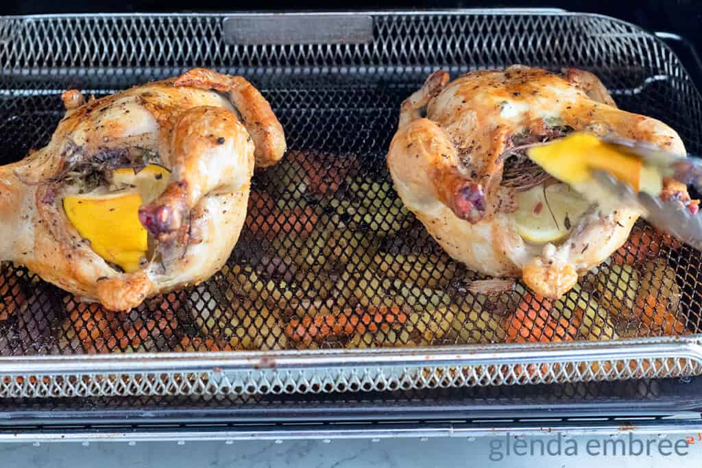 Two roasted Cornish hens in an air fryer with roasted vegetables on the rack underneath them
