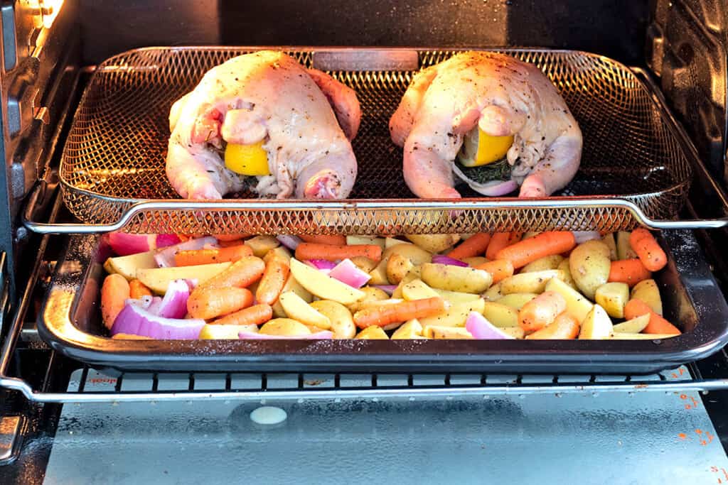 Two raw Cornish hens in an air fryer with a tray of potatoes, carrots and onions on the rack underneath them