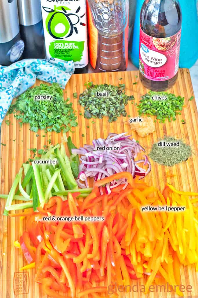 Ingredients for Bell Pepper Salad on a wooden cutting board: julienned bell peppers, red onion, and cucumber with fresh chopped garlic, parsley, basil and chives.