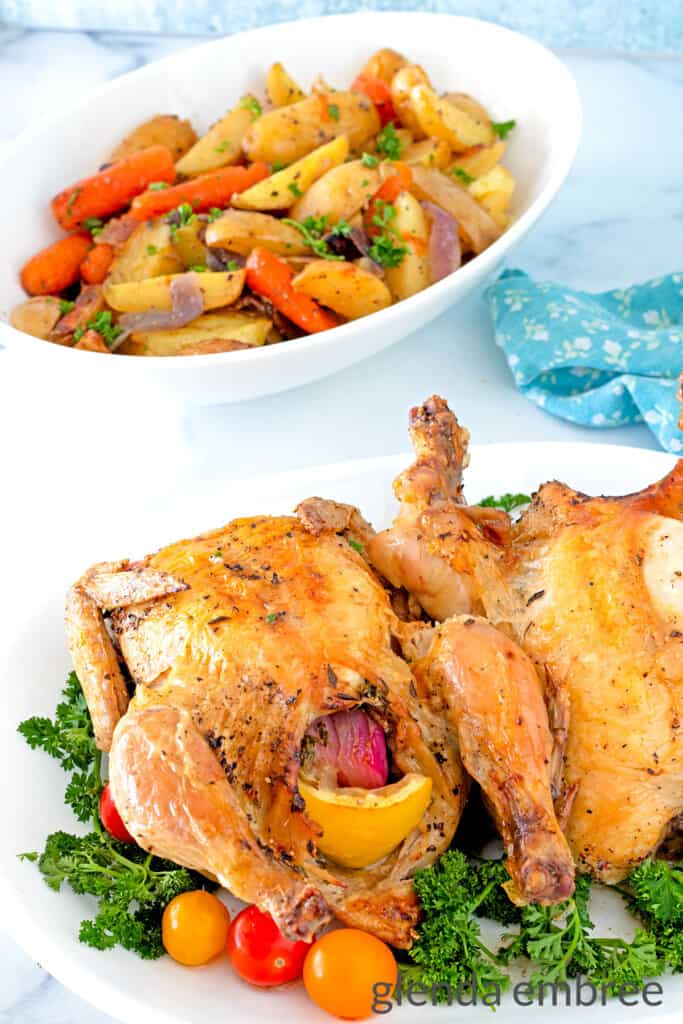 2 Air Fryer Cornish Hen on a white platter garnished with parsley and grape tomatoes.  The platter is in front of a white oval-shaped bowl with roasted potatoes, carrots and onions
