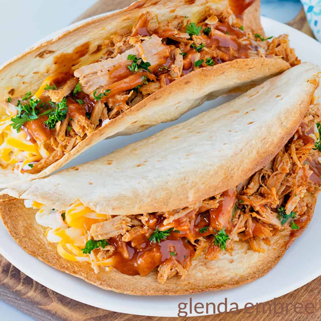 Two Pulled Pork tacos on a white plate with a blue and white print fabric napkin in the background