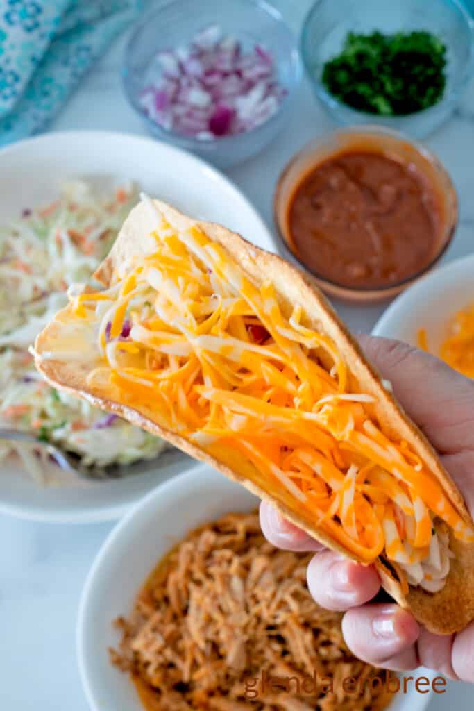 Shredded Colby Jack Cheese added to a crispy flour tortilla shell with pulled pork taco ingredients in the background