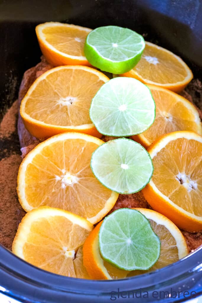 A taco seasoned, raw pork loin in a slow cooker with crisscross pattern cut through the fat cap down to the meat.  Orange and lime slices are laid out across the top of the pork loin.
