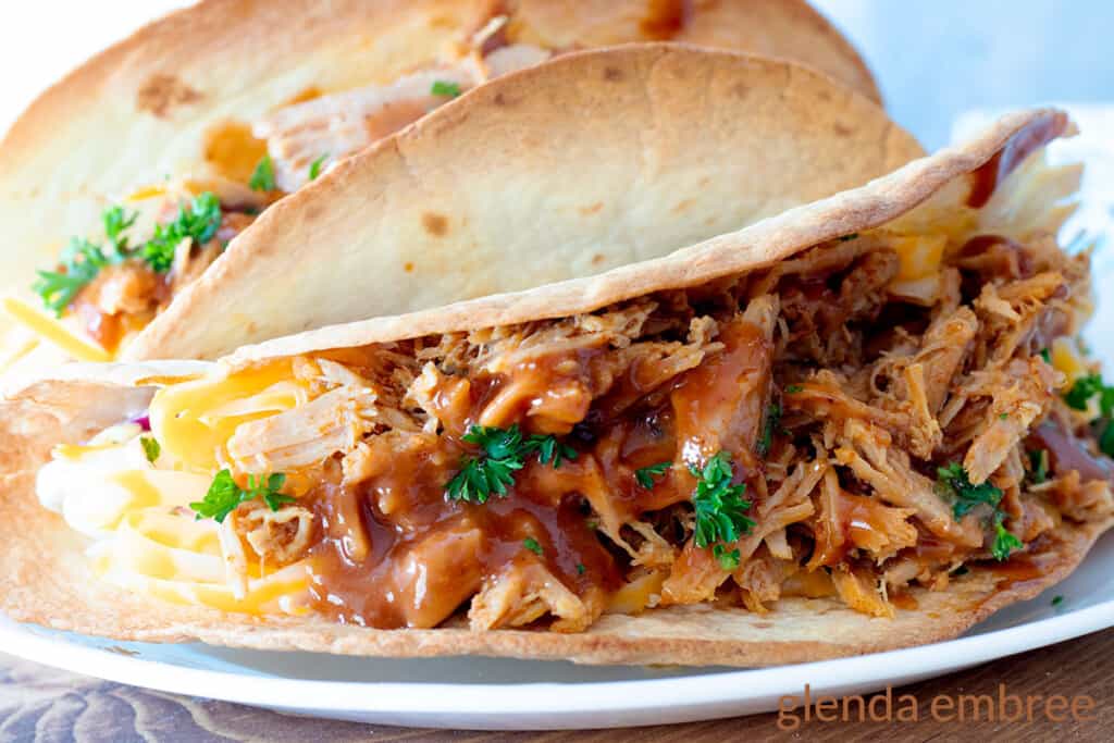 Two Pulled Pork tacos on a white plate with a blue and white print fabric napkin in the background