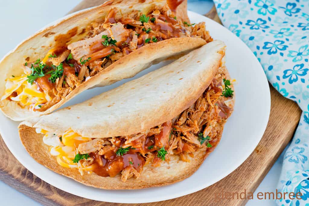 Two Pulled Pork tacos on a white plate with a blue and white print fabric napkin in the background