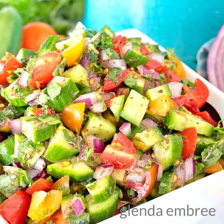 Shirazi Salad in a square, white ceramic bowl on a marble countertop with cucumbers, onions and tomatoes scattered about and a turquoise pitcher in the background