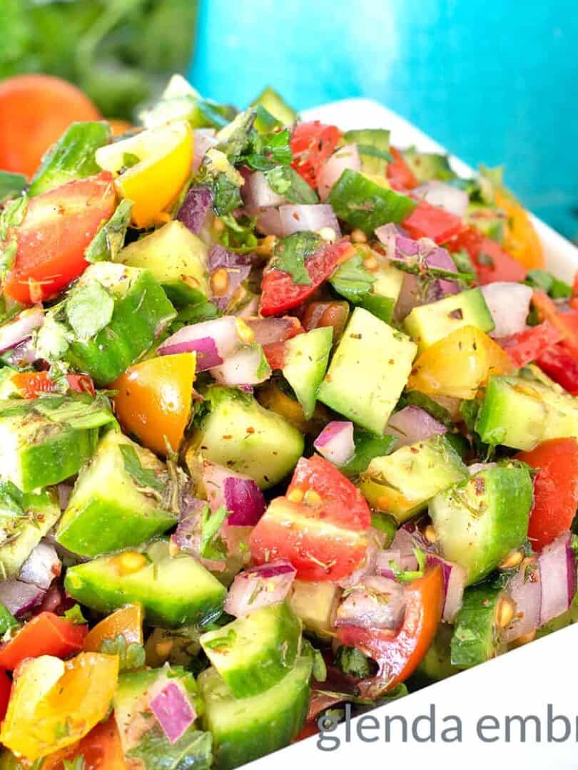 Shirazi Salad in a square, white ceramic bowl on a marble countertop with cucumbers, onions and tomatoes scattered about and a turquoise pitcher in the background
