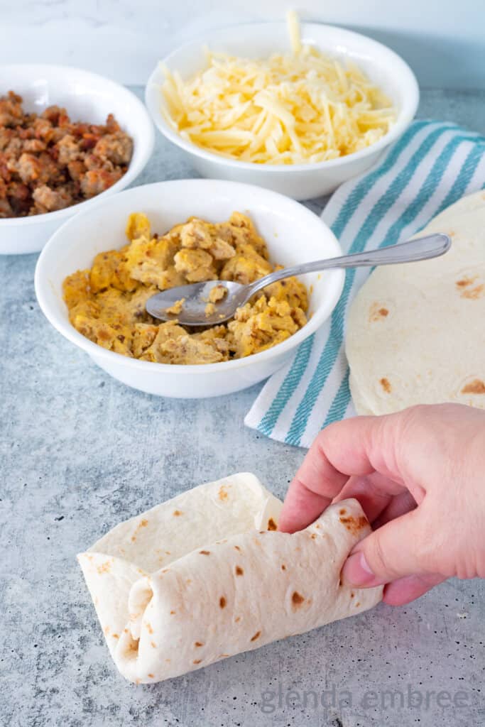 Breakfast Taco ingredients in white bowls, browned sausage, scrambled eggs and grated white Cheddar cheese.  Hand rolling a taco on concrete counter is in the foreground.