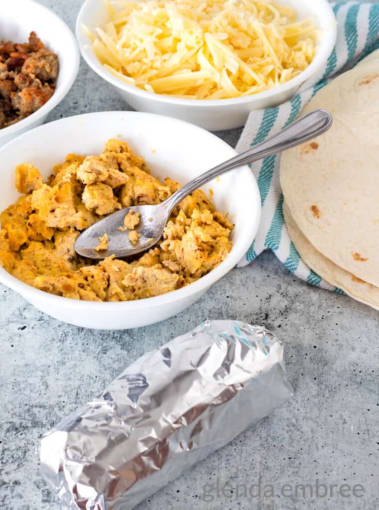 Breakfast taco wrapped in foil on concrete counter.  Bowls with eggs, sausage and grated cheese are in the background next to flour tortillas