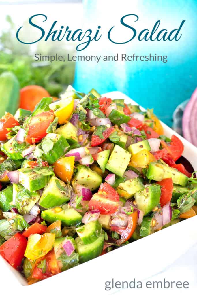 Shirazi Salad in a square, white ceramic bowl on a marble countertop with cucumbers, onions and tomatoes scattered about and a turquoise pitcher in the background