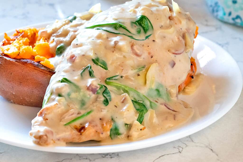 Spinach Artichoke Chicken on a white plate with baked sweet potato.