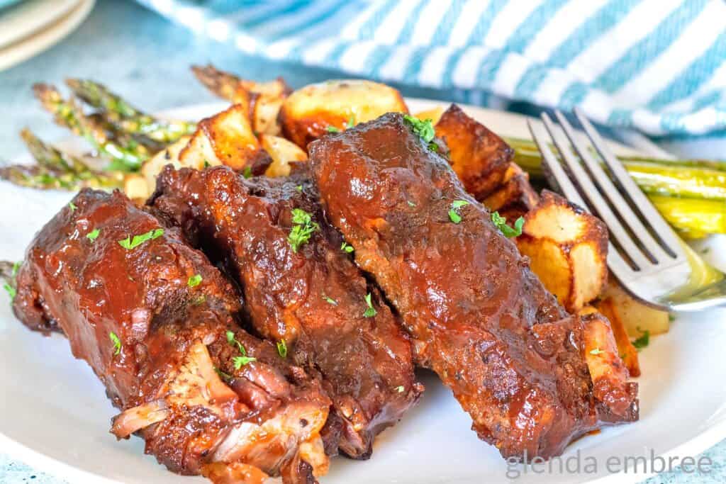 Country Style Ribs on a white plate with roasted potatoes and asparagus.