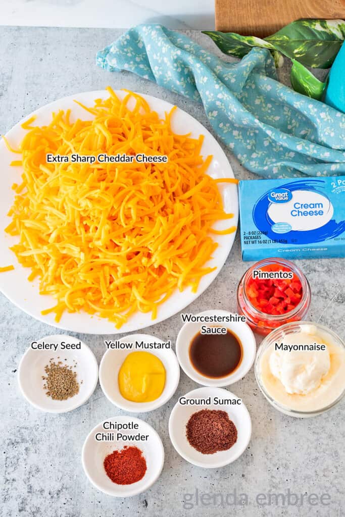 Pimento Cheese ingredients arranged on a countertop: grated Cheddar, cream cheese, mayonnaise, pimentos, Worcestershire sauce, yellow mustard, celery seed, ground sumac and chipotle chili powder.