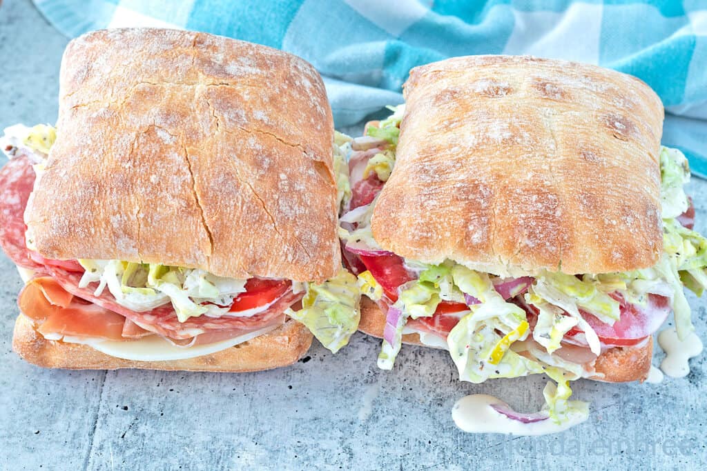 Two Italian grinder sandwiches on ciabatta rolls on a concrete countertop.