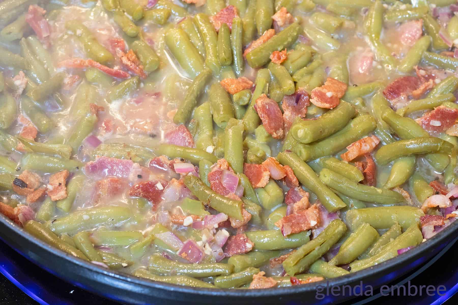 Green Beans simmering in a skillet with minced onion and bacon.