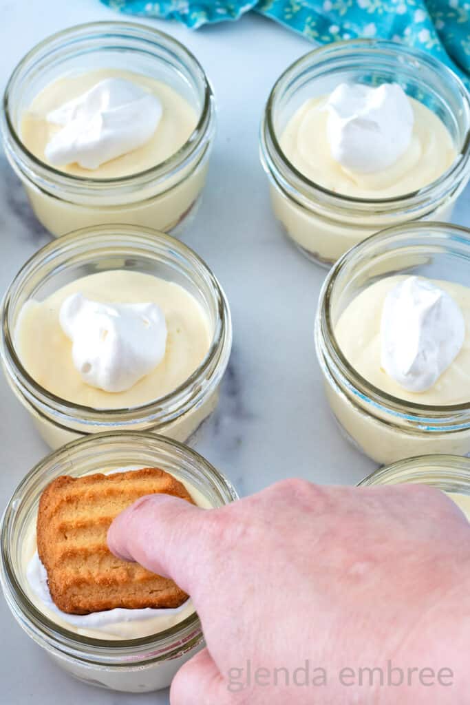 Clear glass half-pint mason jars with a dollop of whipped cream over pudding and a cookie being pressed over the whipped cream.