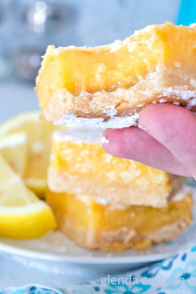Lemon Bar with a bite out of it being held up over a plate of lemon bars.