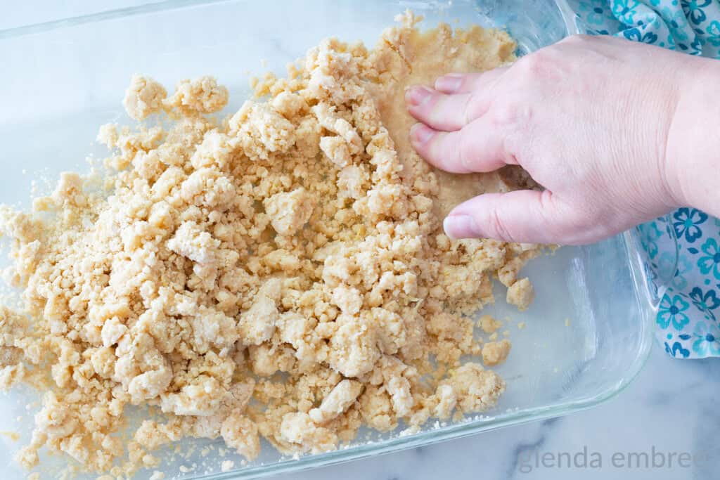 Crumbled shortbread dough being pressed into a 9x12 glass baking pan to form a crust for lemon bars.