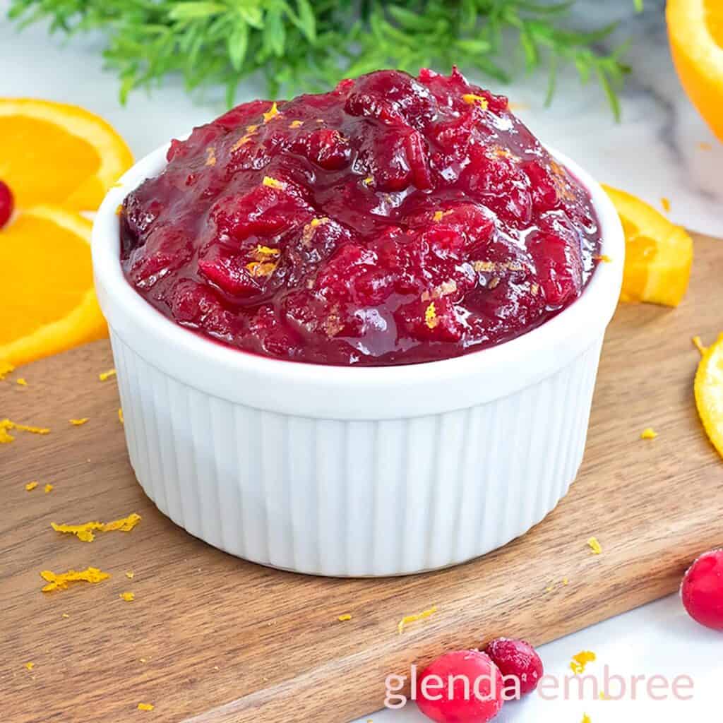 Whole berry cranberry sauce in a white ramekin on a wooden cutting board with orange slices and raw cranberries.