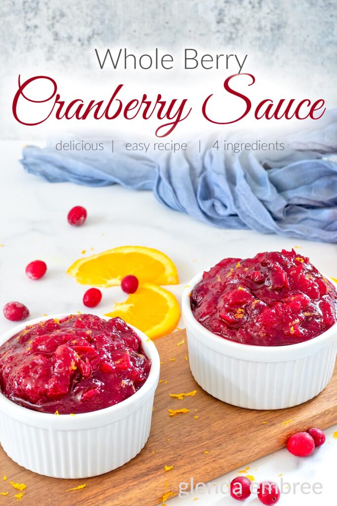 Whole beery cranberry sauce in a white ramekin on a wooden cutting board with orange slices and raw cranberries.