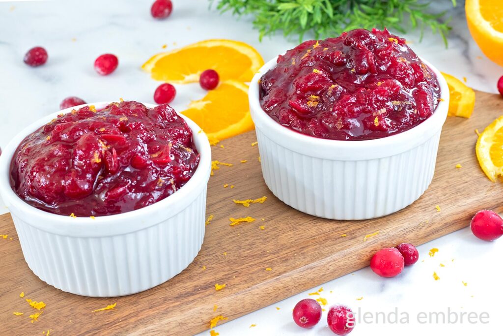 Whole beery cranberry sauce in a white ramekin on a wooden cutting board with orange slices and raw cranberries.