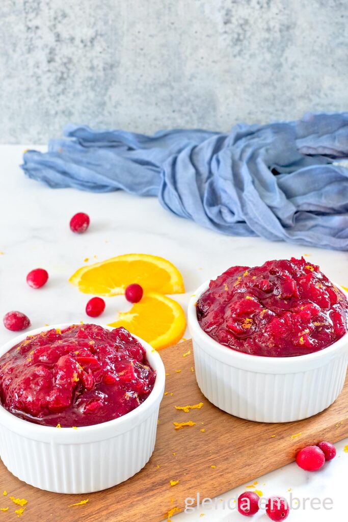 Whole berry cranberry sauce in a white ramekin on a wooden cutting board with orange slices and raw cranberries.