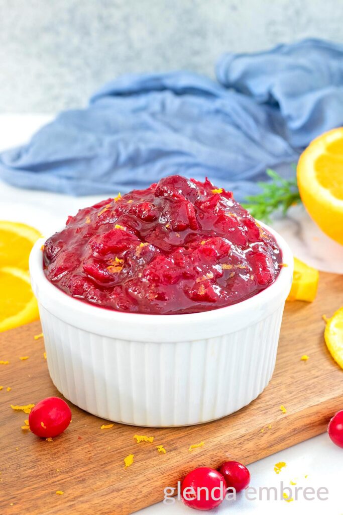 Whole beery cranberry sauce in a white ramekin on a wooden cutting board with orange slices and raw cranberries.