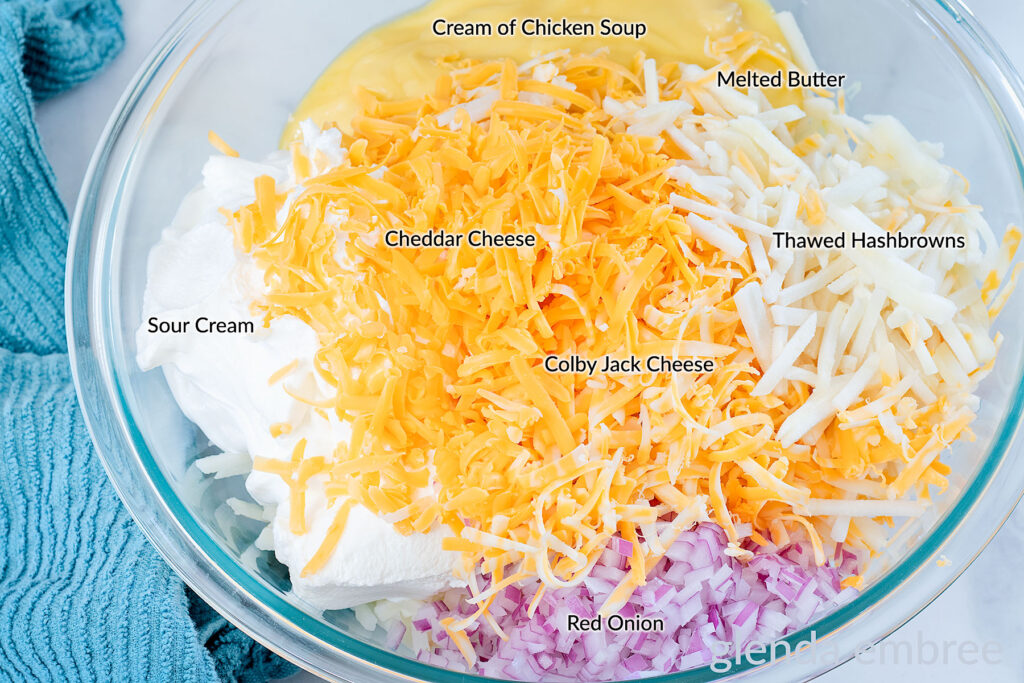 Ingredients for hashbrown casserole in a clear glass mixing bowl — thawed hashbrowns, cream of chicken soup, s our cream, minced red onion, melted butter, Cheddar cheese and Colby Jack cheese.