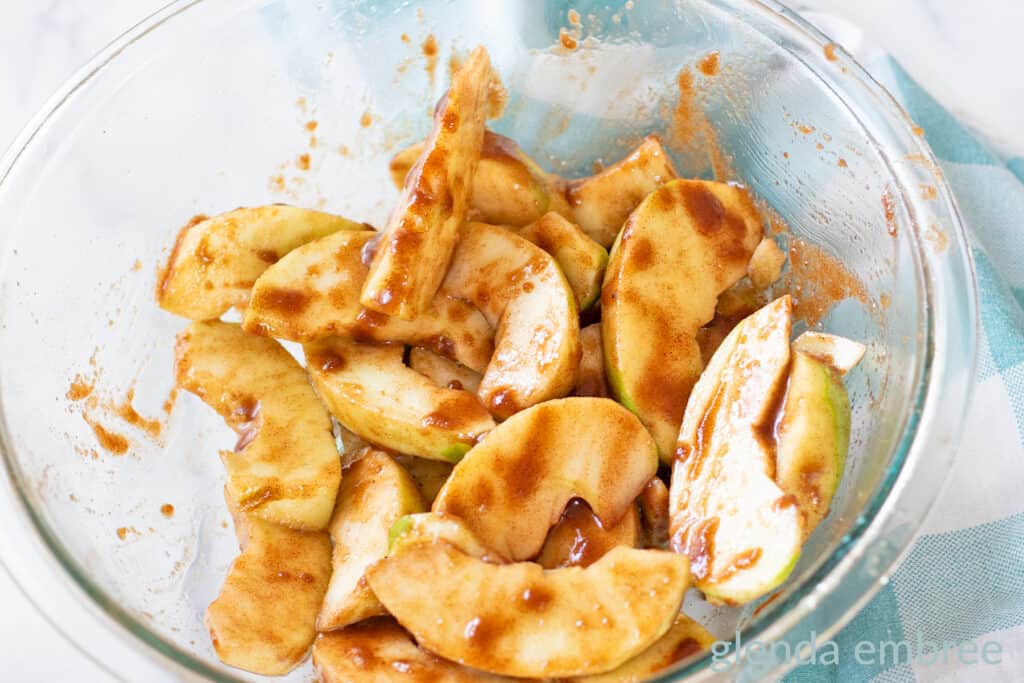 A clear glass bowl with Granny Smith apples that have been peeled, cored and sliced, then tossed with melted butter, brown sugar and cinnamon.