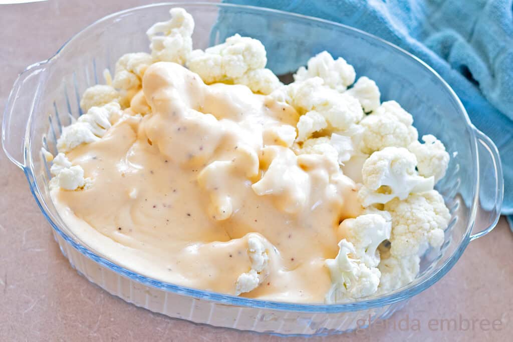 Raw cauliflower florets with cheese sauce poured over in a clear glass baking dish .