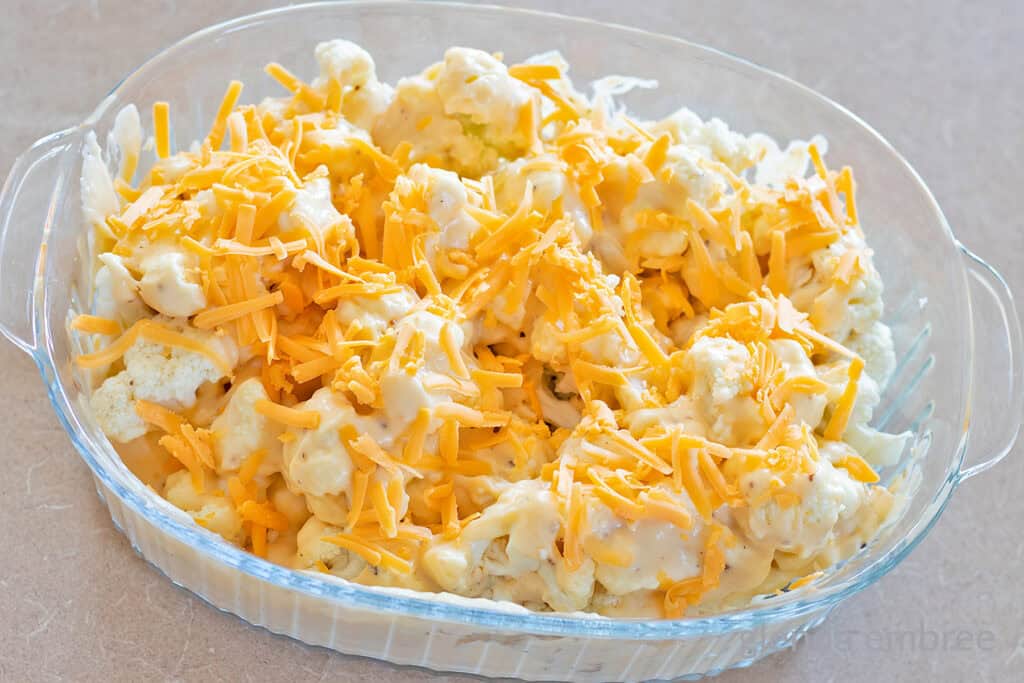 Raw cauliflower florets mixed with cheese sauce and topped with grated cheddar in a clear glass baking dish.