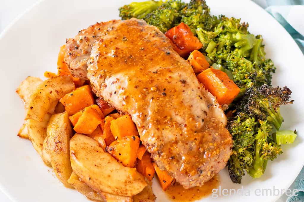 Sheet Pan Honey Mustard Pork Chops on a white plate with roasted sweet potatoes, cinnamon apples and broccoli.