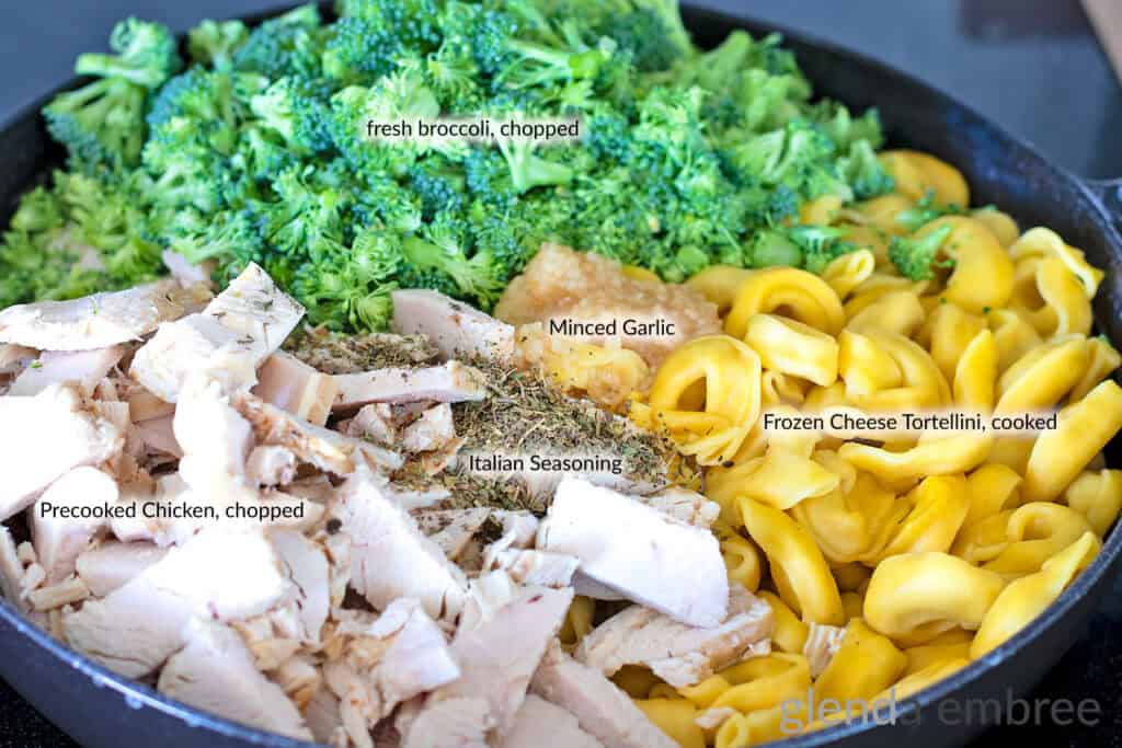 Fresh Broccoli, cooked tortellini, chopped pre-cooked chicken, minced garlic and Italian seasoning in a cast iron skillet in preparation for making Creamy Chicken Tortellini Bake.