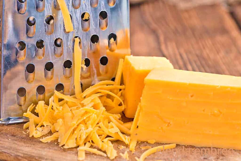 Cheddar Cheese being grated on a wooden cutting board with a box grater.