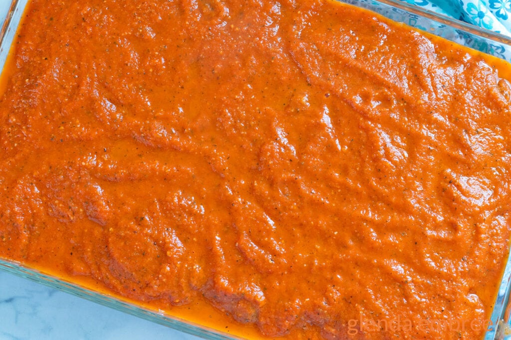 Marinara sauce poured over raw meatballs in a clear glass baking dsih.