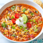 Taco Soup in a white ceramic bowl, garnished with grated cheese, sour cream, tomatoes, green onion and a lime wedge.