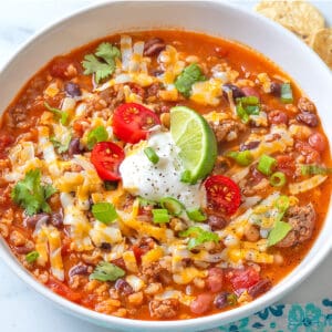 Taco Soup in a white ceramic bowl, garnished with grated cheese, sour cream, tomatoes, green onion and a lime wedge.