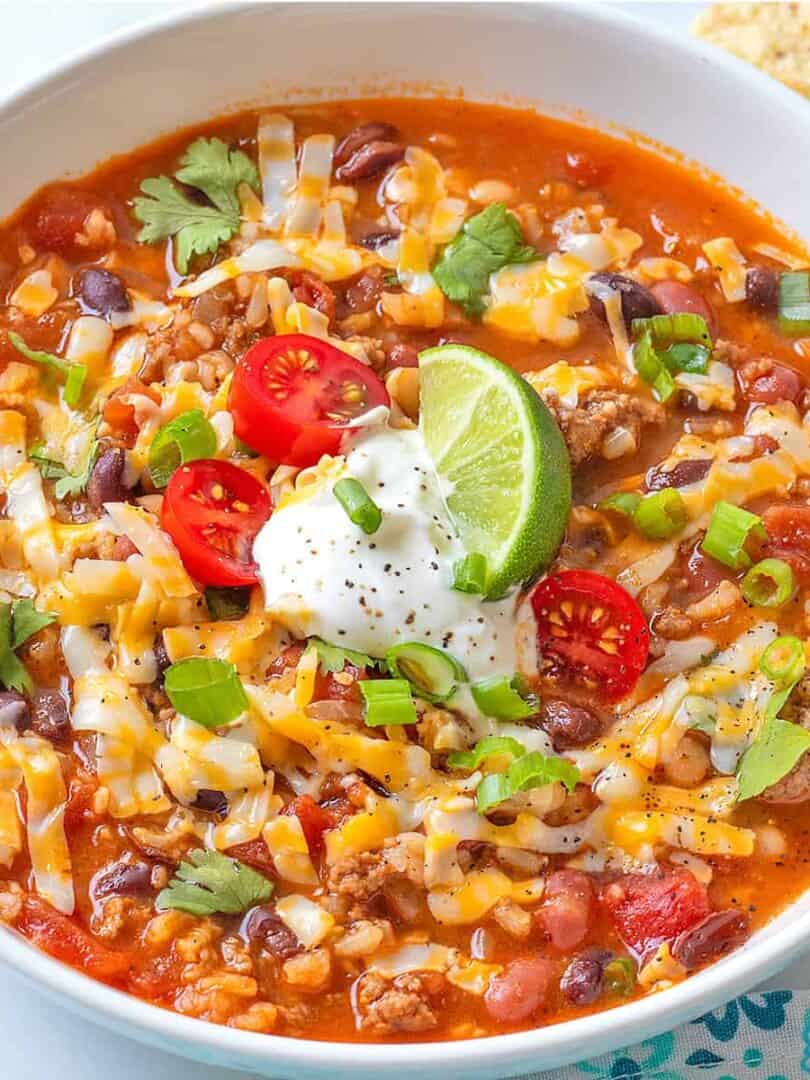Taco Soup in a white ceramic bowl, garnished with grated cheese, sour cream, tomatoes, green onion and a lime wedge.