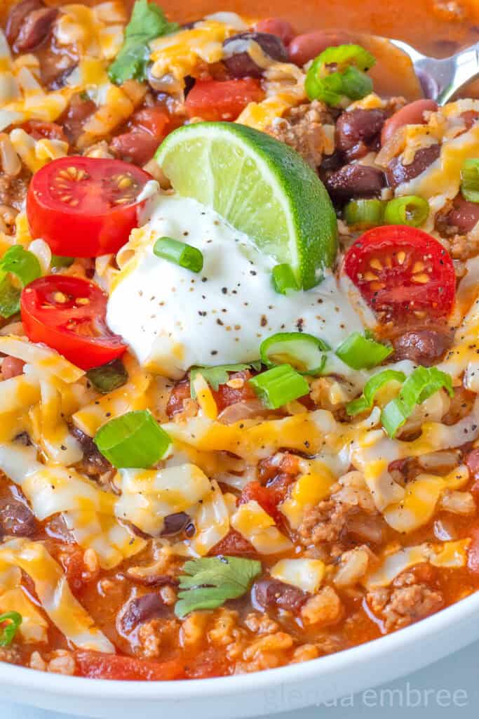Taco Soup in a white ceramic bowl, garnished with grated cheese, sour cream, tomatoes, green onion and a lime wedge.