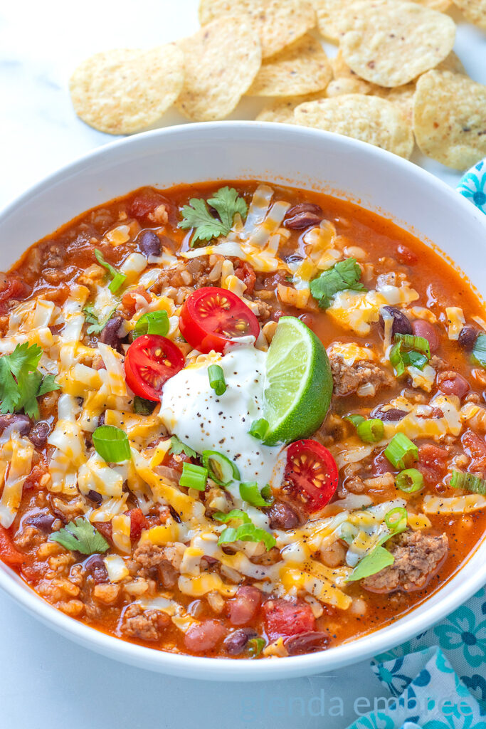 Taco Soup in a white ceramic bowl, garnished with grated cheese, sour cream, tomatoes, green onion and a lime wedge.