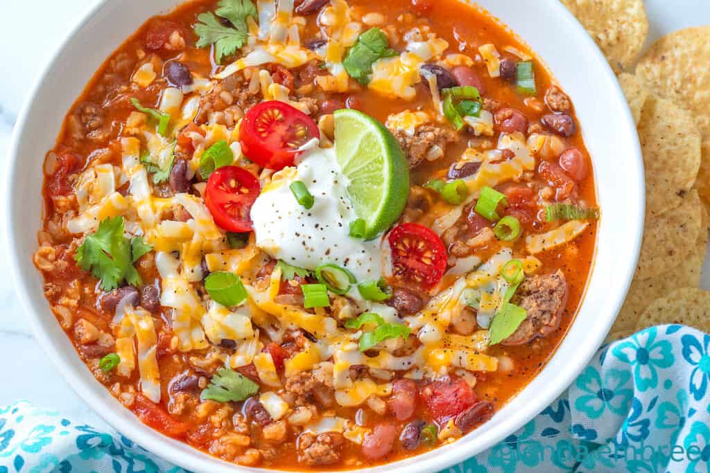 Taco Soup in a white ceramic bowl, garnished with grated cheese, sour cream, tomatoes, green onion and a lime wedge.