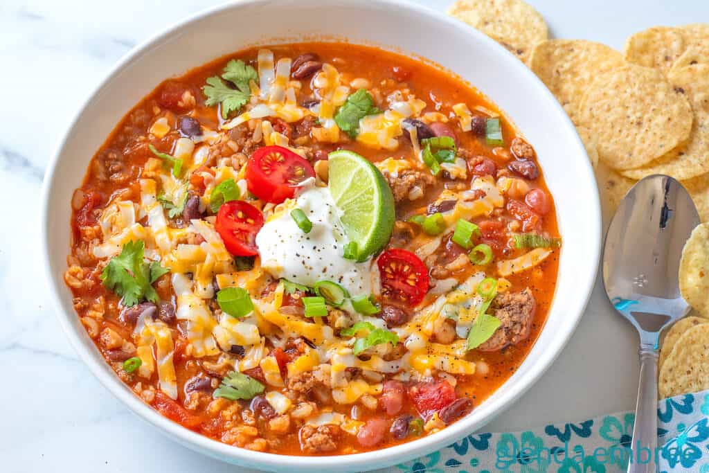 Taco Soup in a white ceramic bowl, garnished with grated cheese, sour cream, tomatoes, green onion and a lime wedge.