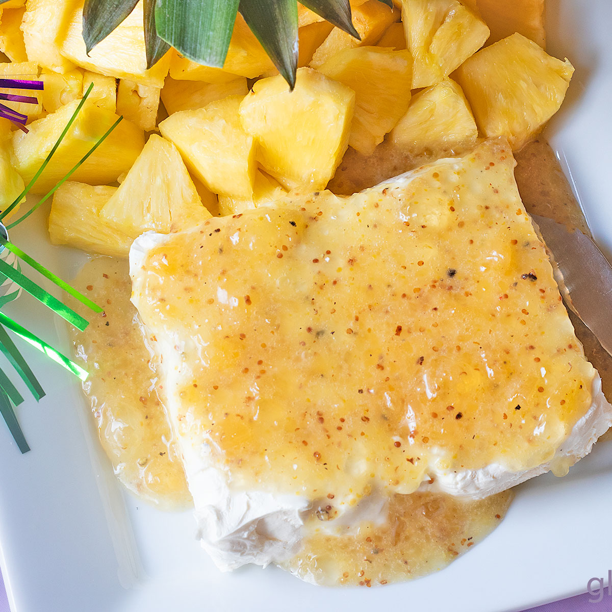 Jezebel Sauce over Cream Cheese on a white rectangular stoneware platter with a pineapple stem and fresh pineapple chunks.