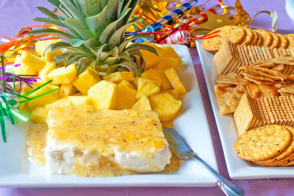 Jezebel Sauce over Cream Cheese on a white rectangular stoneware platter with a pineapple stem and fresh pineapple chunks.