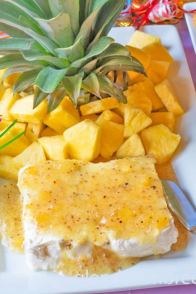 Jezebel Sauce over Cream Cheese on a white rectangular stoneware platter with a pineapple stem and fresh pineapple chunks.