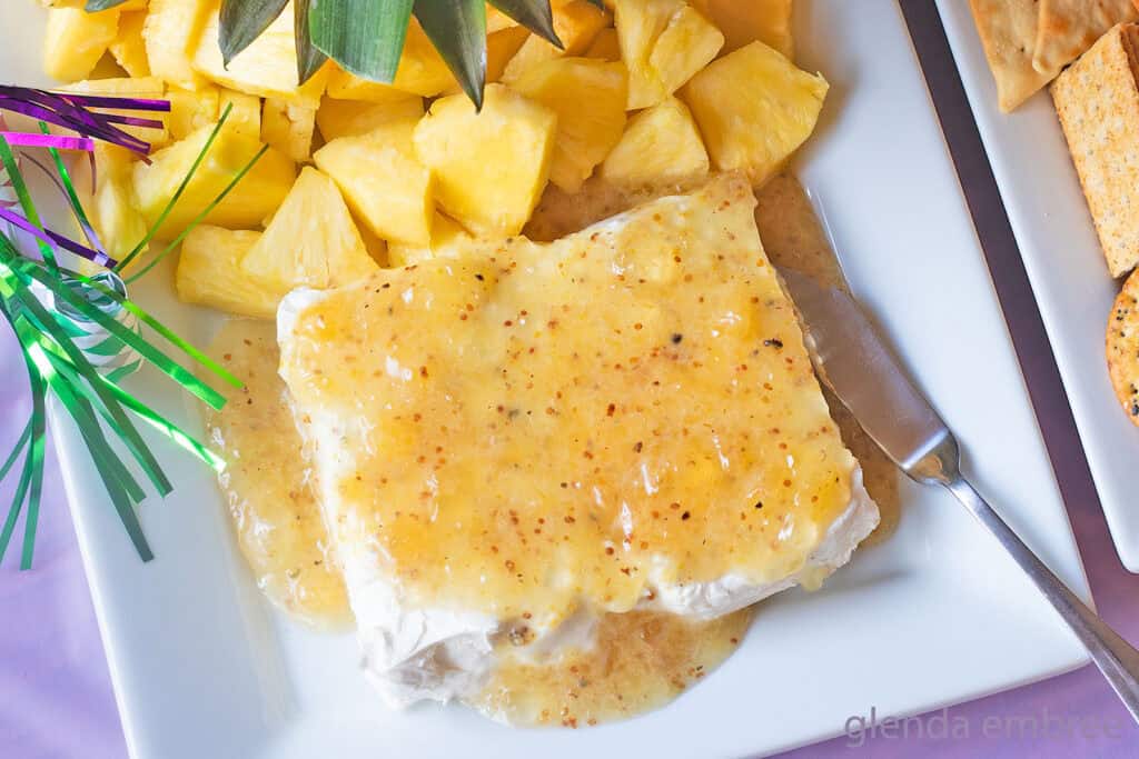 Jezebel Sauce over Cream Cheese on a white rectangular stoneware platter with a pineapple stem and fresh pineapple chunks.
