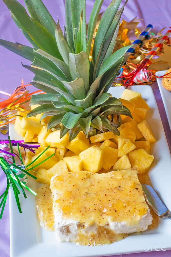 Jezebel Sauce over Cream Cheese on a white rectangular stoneware platter with a pineapple stem and fresh pineapple chunks.